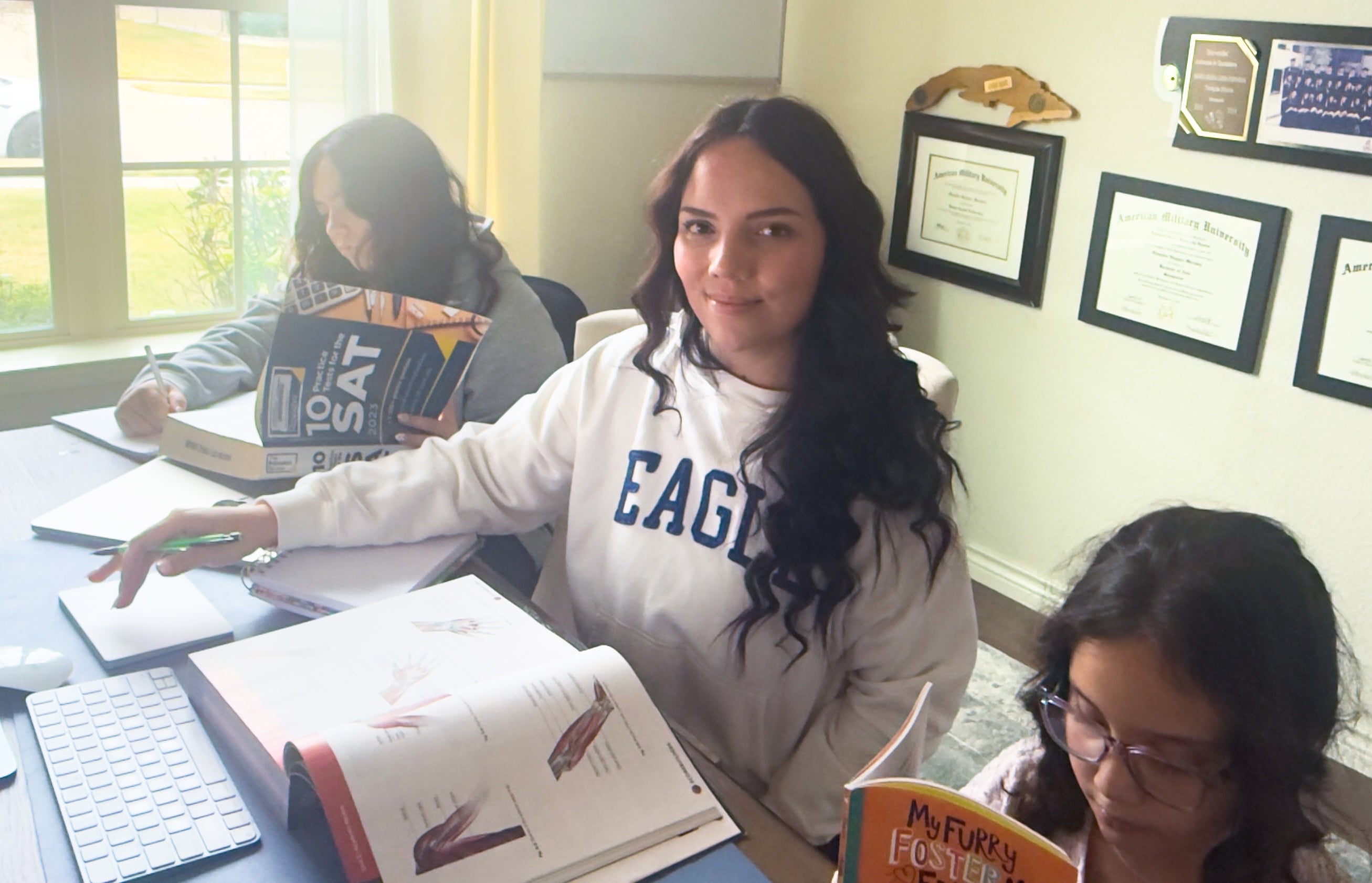 A woman with long dark hair sits at a desk with two small children next to her