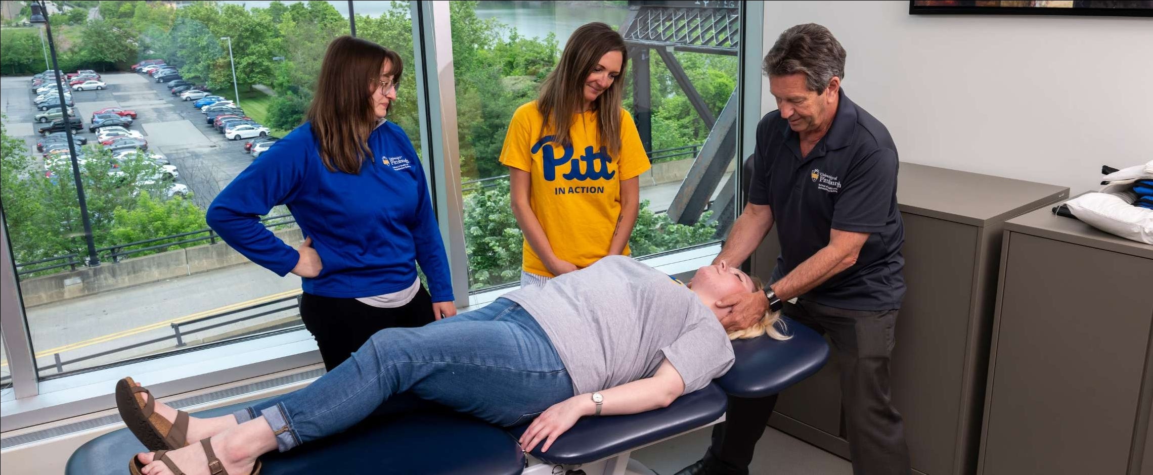 Students observing a patient model on a medical table while professor is demonstrating an adjustment.