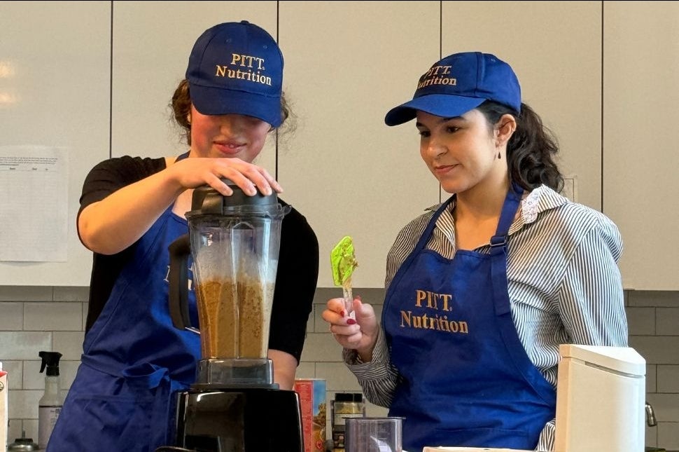 Two students using a blender wearing Pitt Nutrition Aprons