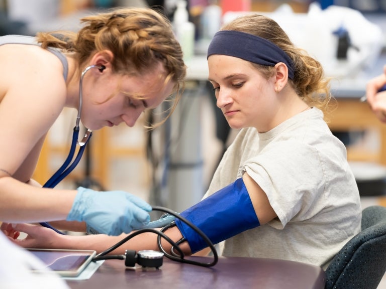 Two students in a lab classroom, one on the left has her hair braided and is using gloves and a blood pressure machine on the girl on the right who is wearing a white shirt and a navy blue headband.