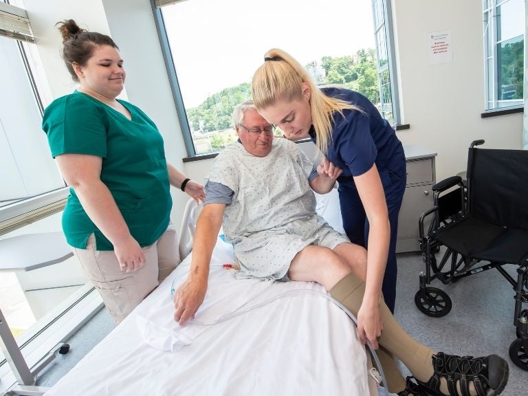 Students working with an elderly model patient on a hospital bed.