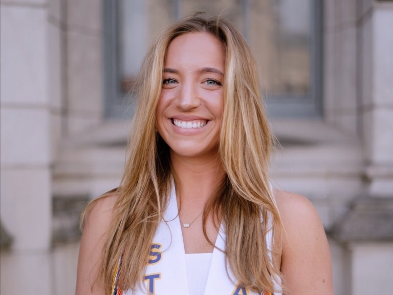 Woman with blonde hair wearing her graduation sash in front of a Pitt building.