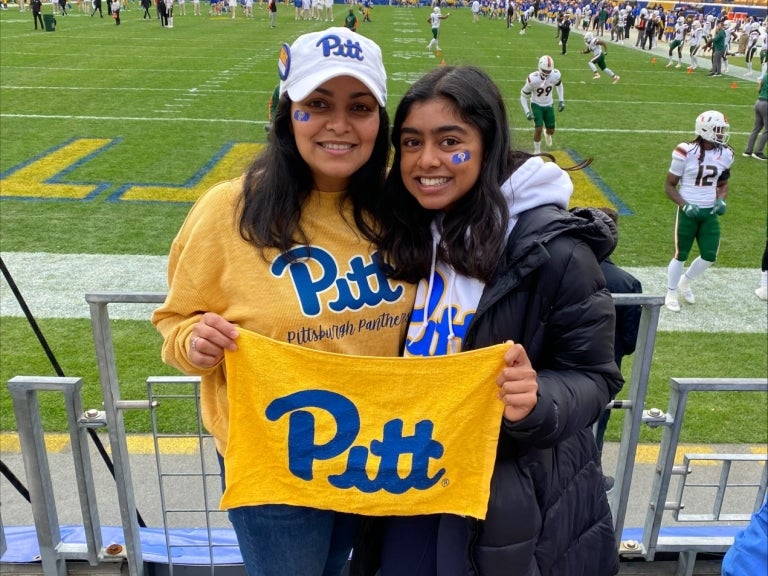 Two brunette women standing next to each other wearing University of Pittsburgh shirts and hats and holding a yellow Pitt towel in front of a football field. 