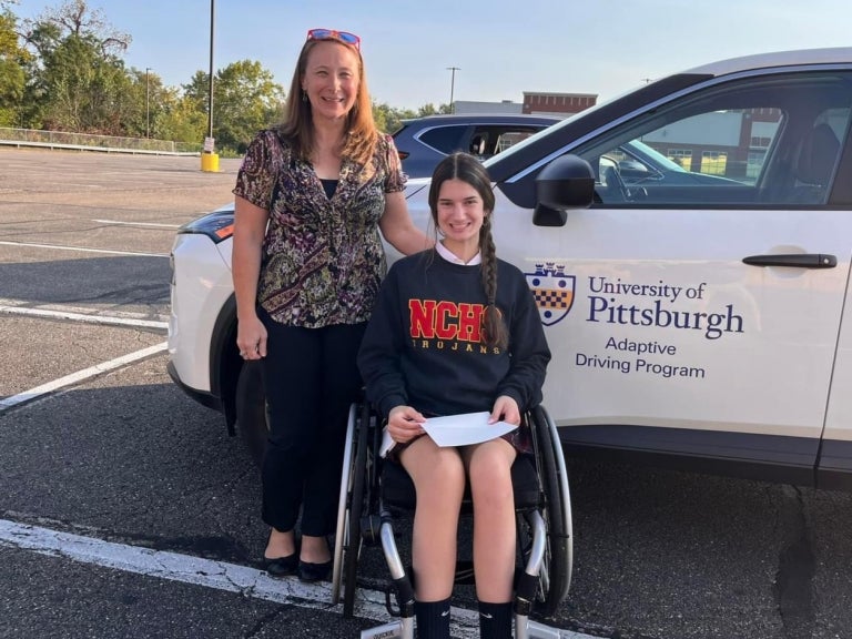 Melissa Aleksak stands next to her client Mary who sits in a wheelchair smiling while holding her graduation certificate. Behind them is a white car with University of Pittsburgh Adaptive Driving Program written on the driver's door