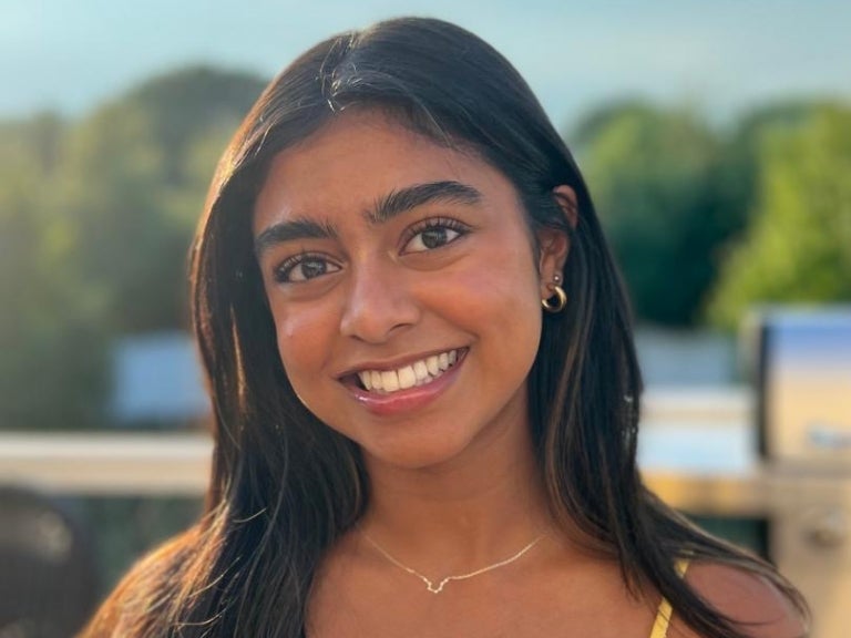 A woman with dark brown hair wearing a yellow top, gold earrings and a gold necklace.