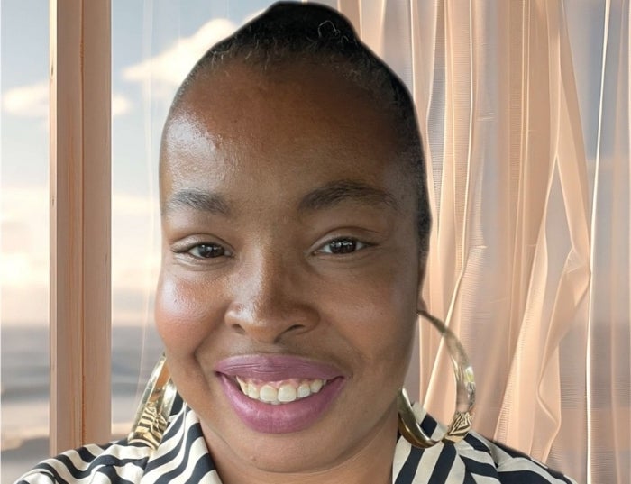 Woman with dark hair wearing gold hoop earrings and a striped collared shirt in front of a window and a beige curtain.