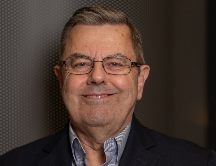 Man in a striped collared shirt and a black suit jacket against a dark gray background.