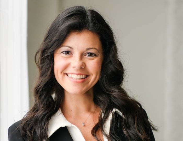 Woman with dark hair in a black blazer over a white shirt in front of a gray background.