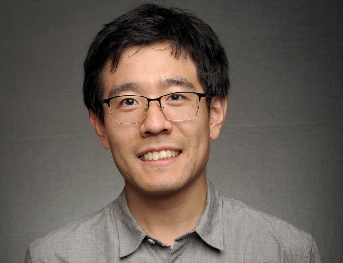 Man with short black hair wearing a gray shirt in front of a dark gray background.