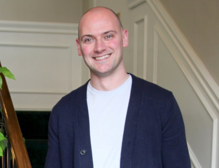 A man wearing a dark cardigan over a white shirt standing on a staircase.