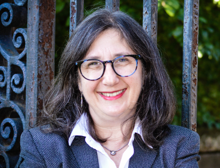 Woman (Sharon McCague) with dark hair and glasses in a blazer and a collared shirt in front of a fence.