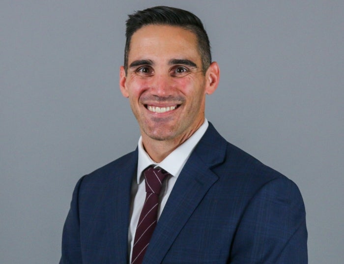 Man with short brown hair and a navy suit in front of a gray background.