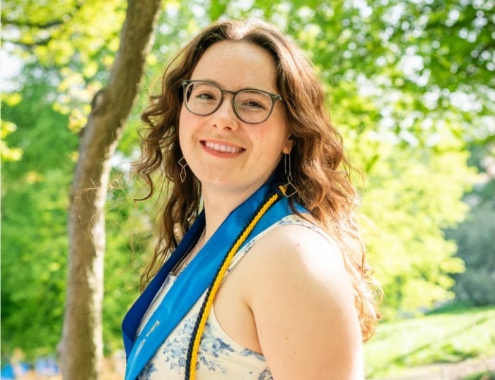 Woman with auburn hair wearing glasses and a white and blue top standing outside in front of a green tree.