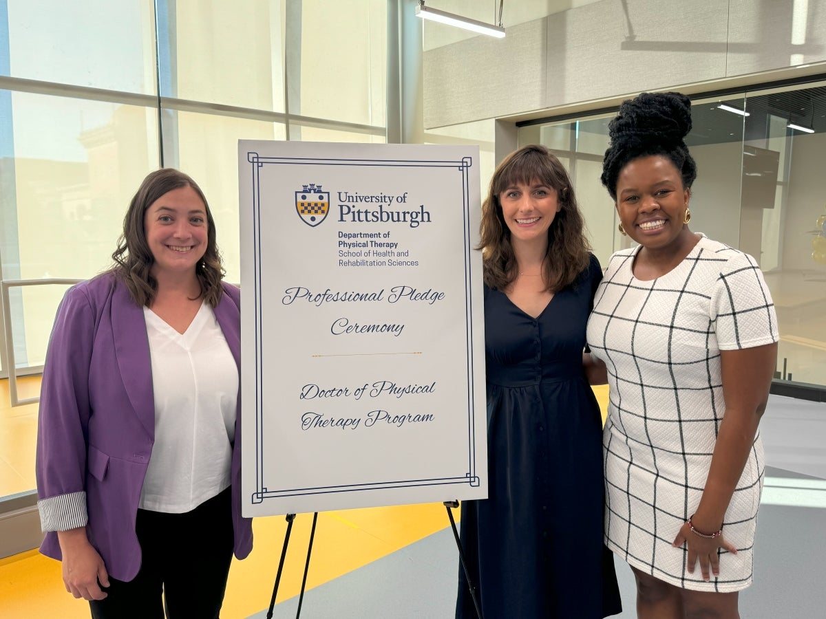 Three women stand smiling next to a sign for the Pitt PT Professional Pledge Ceremony