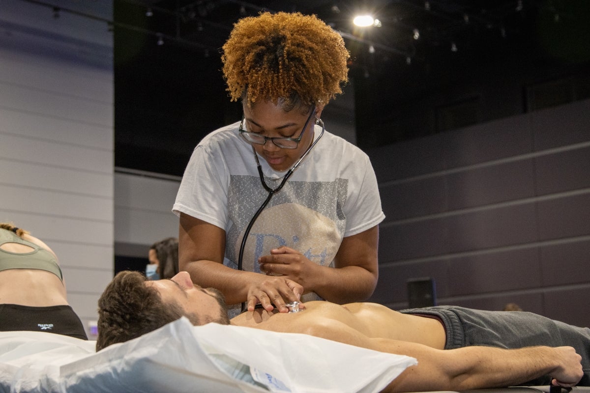 A female student with curly hair stands over a male student laying on a table. She holds a stethoscope to listen to his heartbeat.