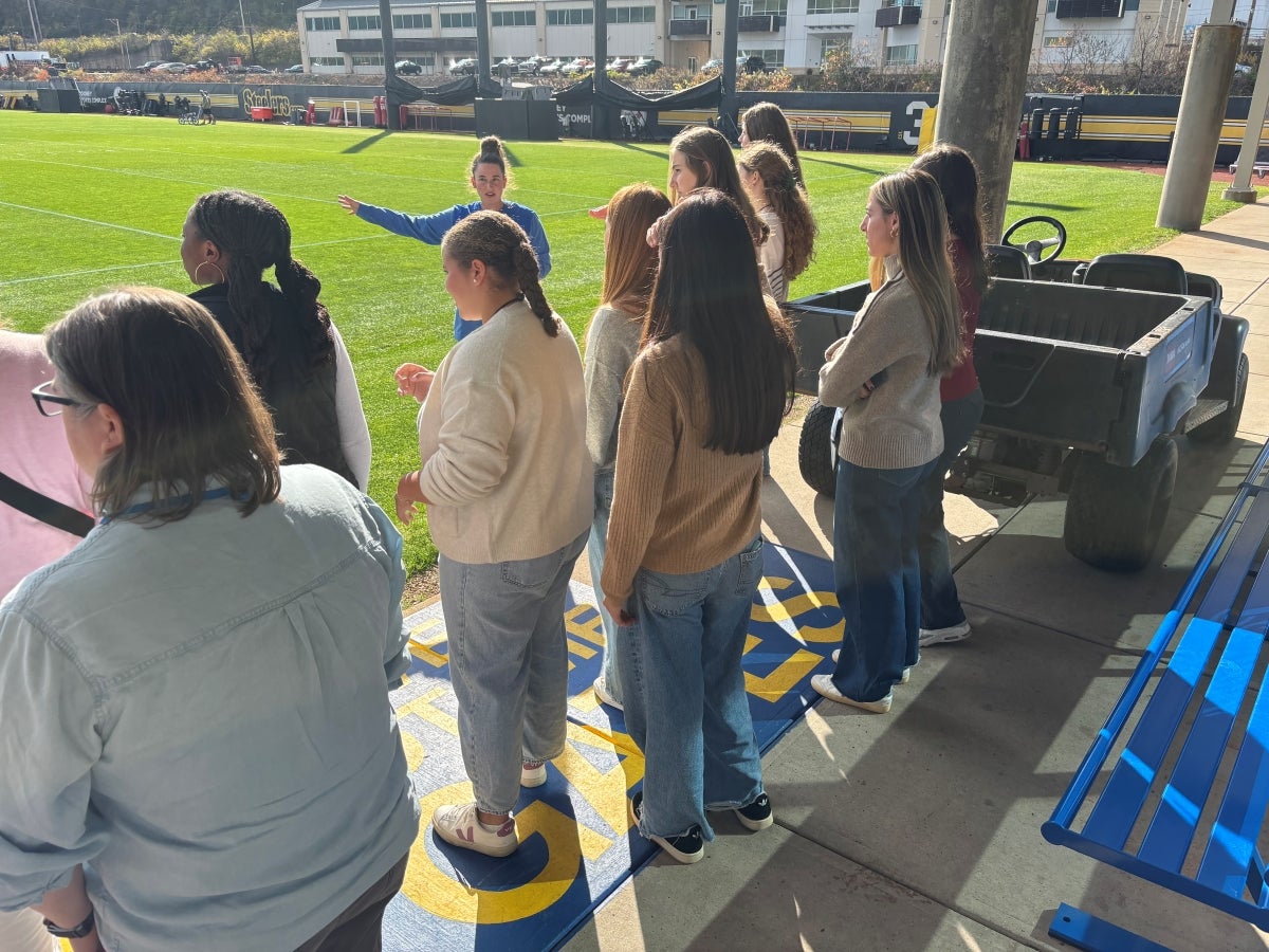 A group of young women stand on a football field listening to an instructor