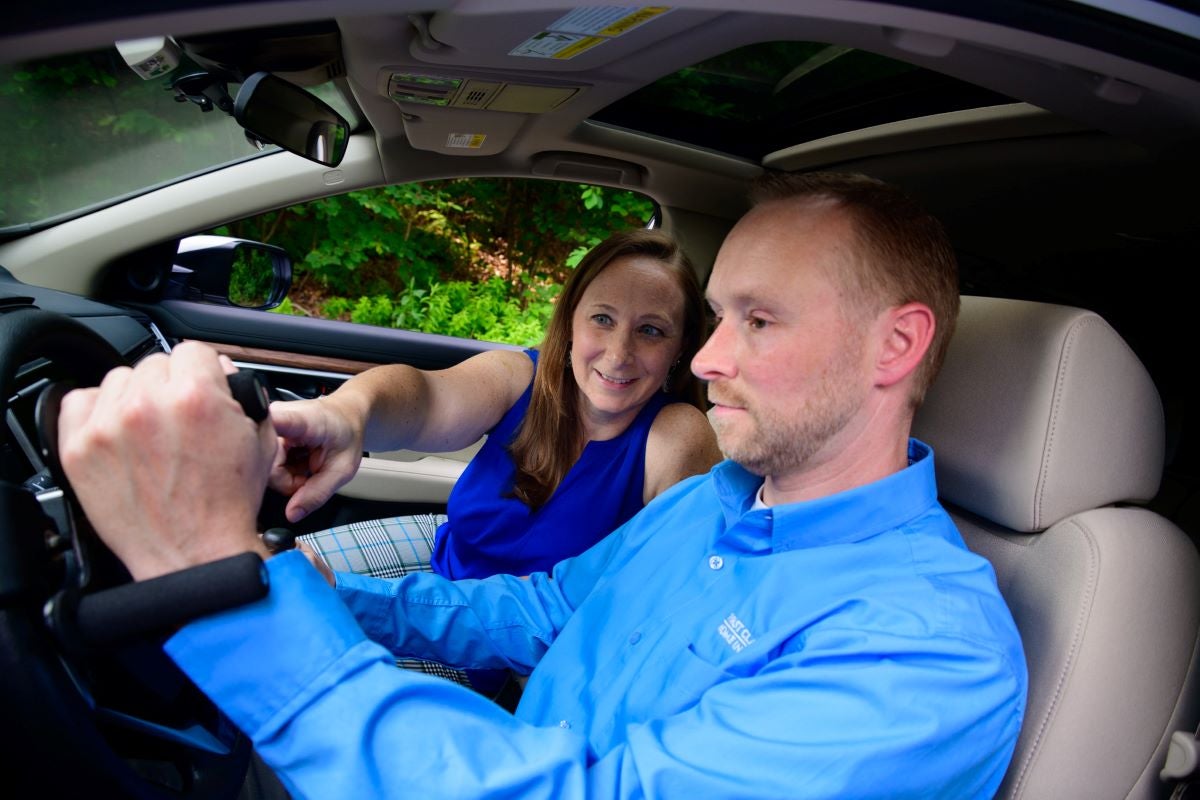 Melissa Aleksak sits in a car's passenger seat while pointing to the stearing wheel with her right hand as she explains to a male driver how to hold the steering wheel's adaptive equipment