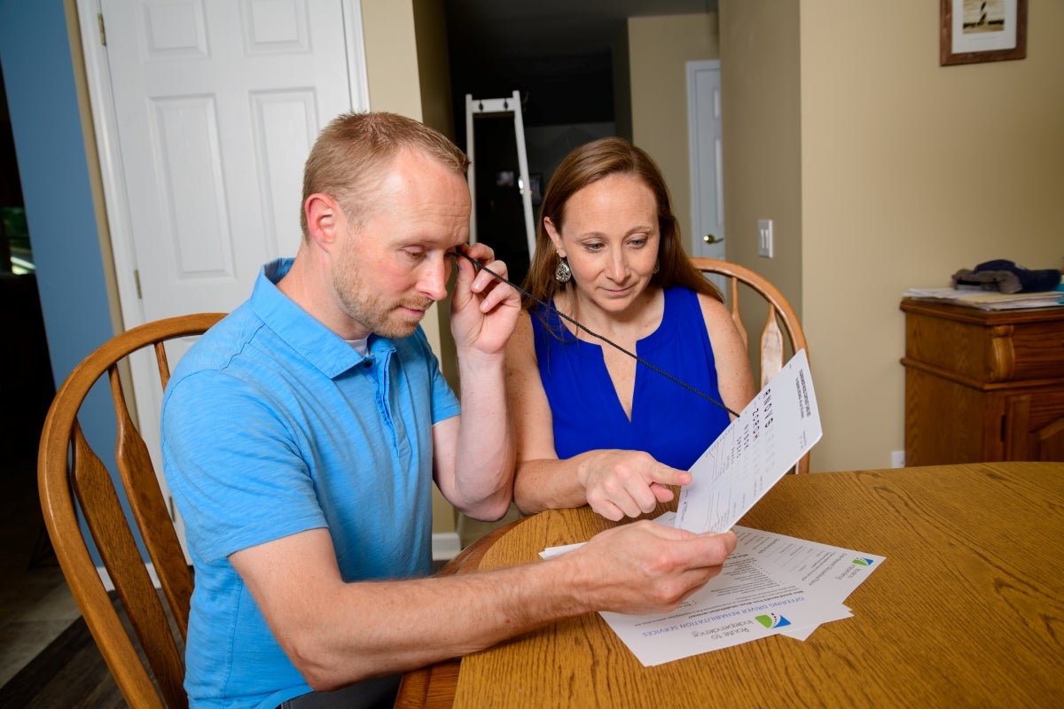 Melissa Aleksak in a dark blue dress sits to the left of a seated male client in a light blue shirt. The client holds a thin black plastic stick between his left eye and a vision chart held by his right hand