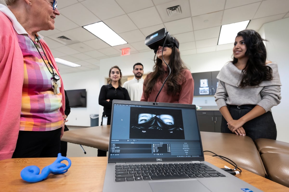 A professor in a pink sweater stands on the left watching a student wearing specialized headgear covering her eyes as data is collected on a laptop computer in the foreground