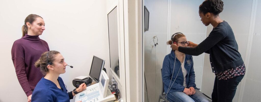 Three professionals working with a patient model in a sound booth.