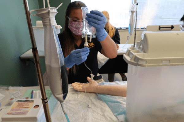 Masked student in a class working with a model arm and IV bag.