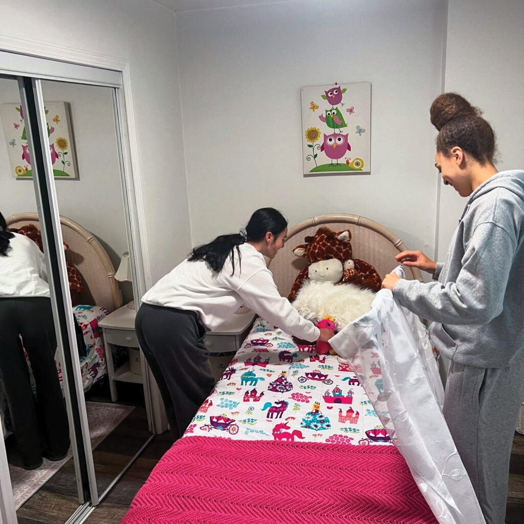Two students making up a bed and setting up curtains on a curtain rod.