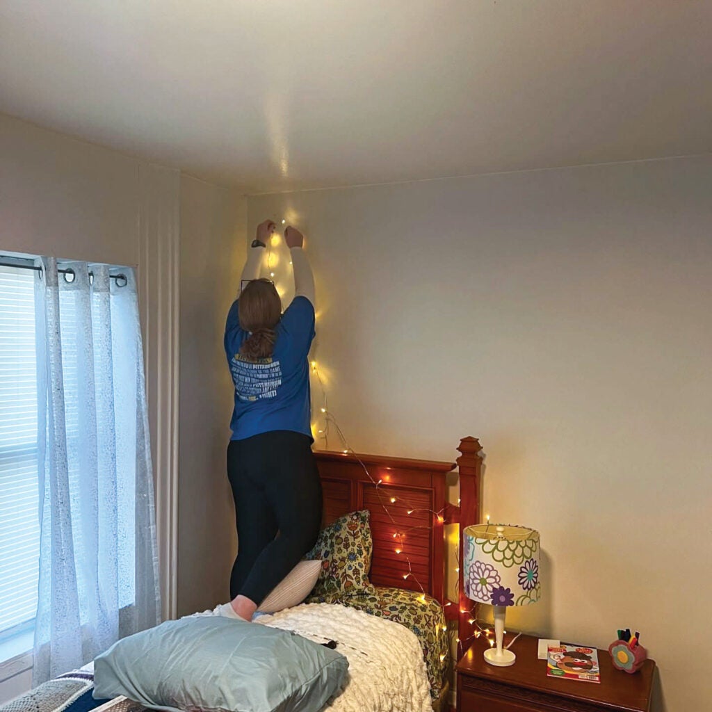 Student hanging up string lights over a twin-sized bed.