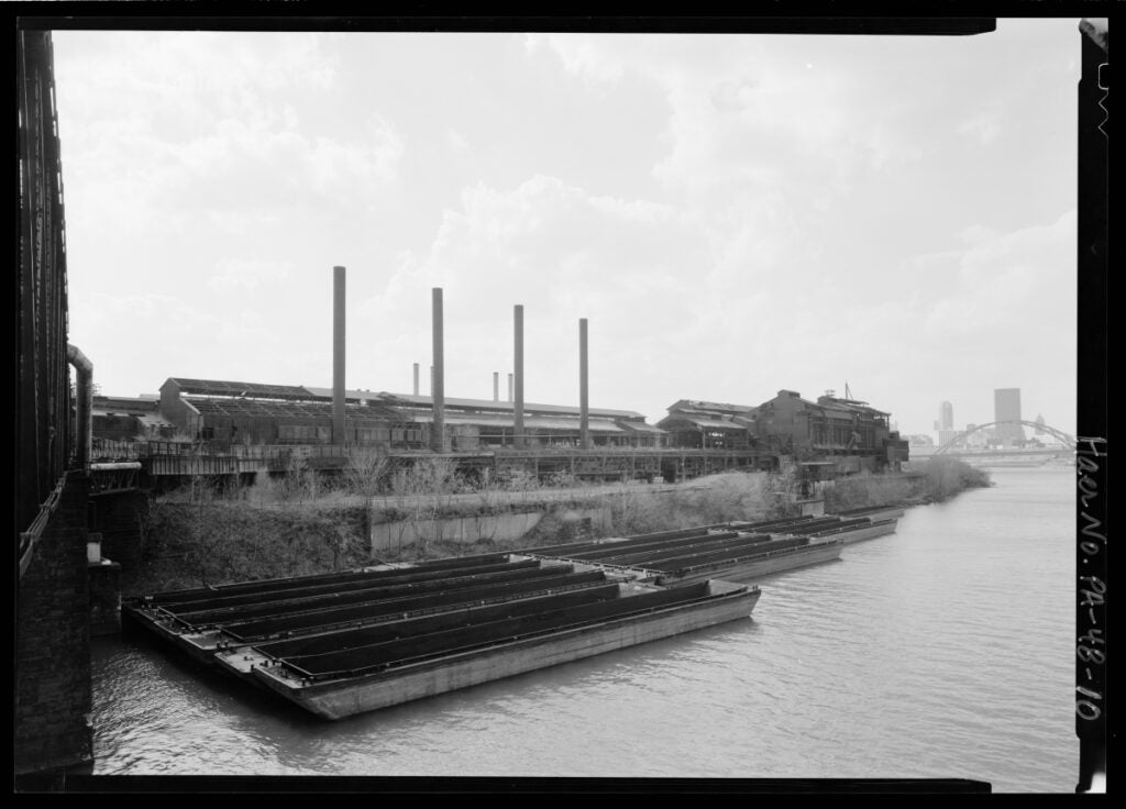 A black and white photo shows a decommissioned steel mill on the banks of a river with the city of Pittsburgh in the background.