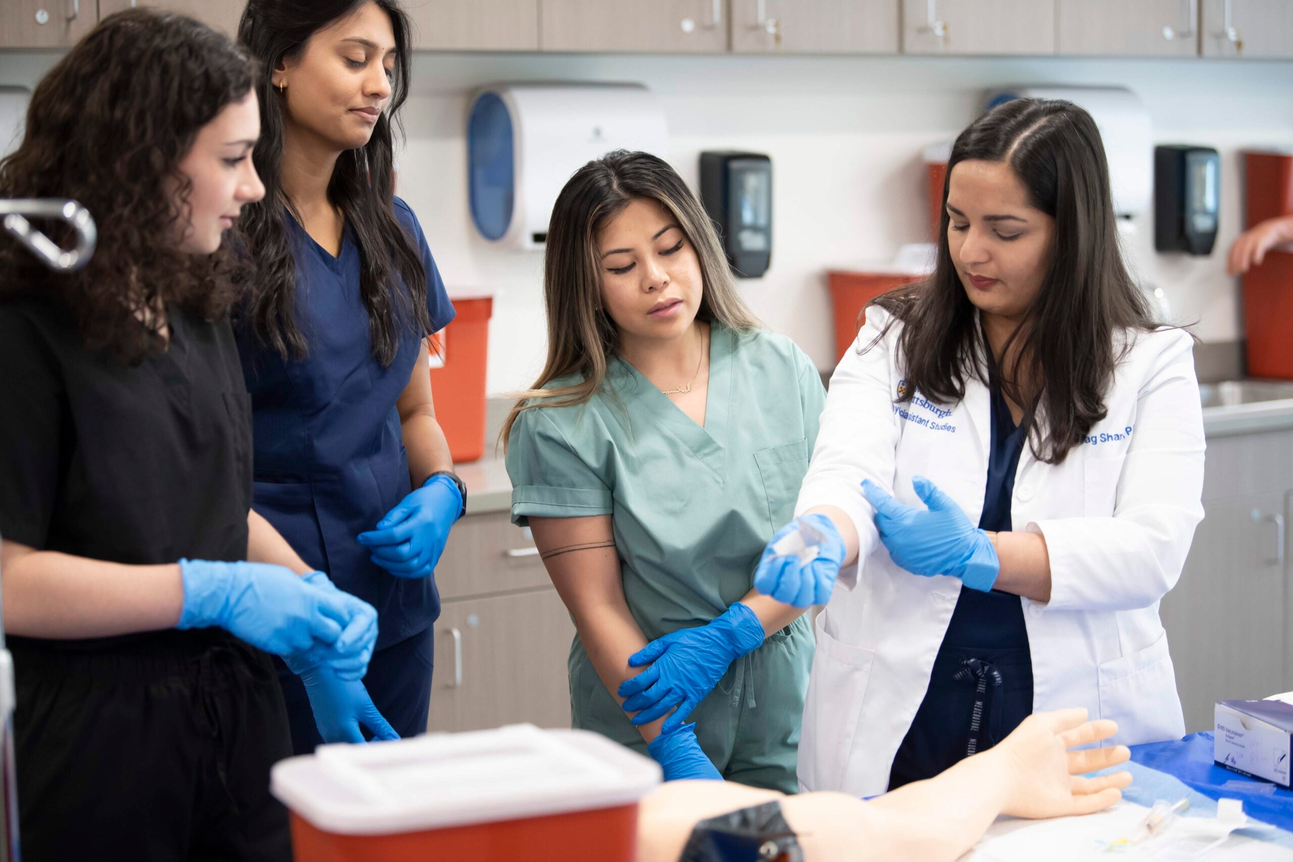 Three female students watching a female instructor in a white coat demonstrate a technique with a syringe.