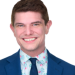 Man with short red hair wearing a blue suit jacket over a light blue and pink floral collared shirt with a dark blue tie.