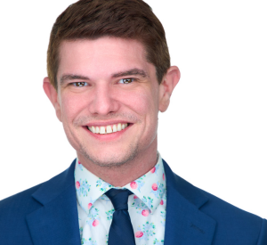 Man with short red hair wearing a blue suit jacket over a light blue and pink floral collared shirt with a dark blue tie.