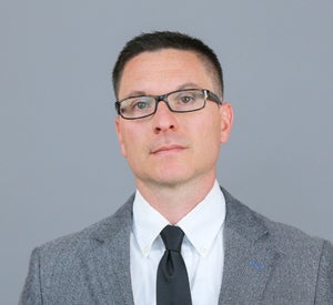 Man with short black hair and glasses wearing a light grey suit and black tie.