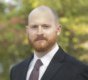 Man with very short hair and a beard wearing a black suit jacket over a white collared shirt and a brown tie.