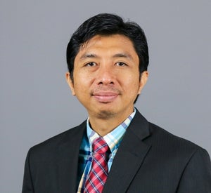 Man with short black hair wearing a black suit jacket over a blue and yellow plaid collared shirt and a red and blue plaid tie.