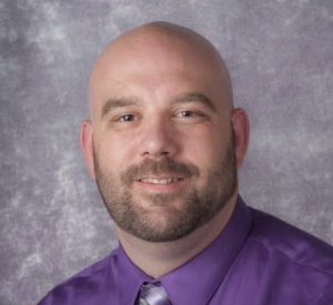 Bald man with a beard wearing a purple collared shirt with a silver tie.