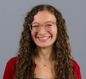 Woman with long, curly brown hair wearing clear glasses, a red sweater and a silver necklace.