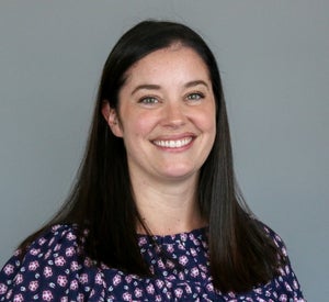 Woman with dark brown hair wearing a purple top with purple and white flowers printed on it.