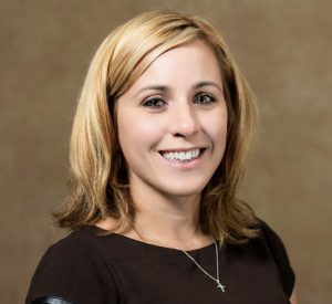 Woman with short blonde hair wearing a dark brown shirt and a silver necklace.