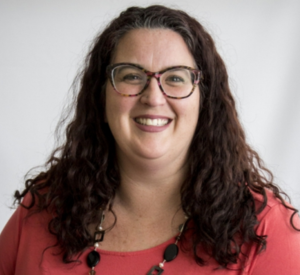 Woman with long brown curly hair wearing a bright red shirt with a brown beaded necklace.