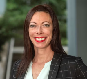 Woman with dark brown hair wearing a black blazer with white lines over a white blouse.