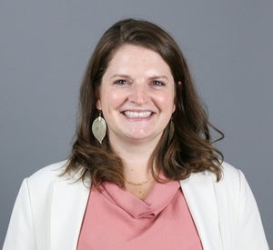 Woman with short brown hair wearing a white blazer over a light pink blouse and gold leaf earrings.