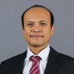 Man with short black hair wearing a black suit jacket over a white collared shirt and a red and blue plaid tie.