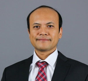 Man with short black hair wearing a black suit jacket over a white collared shirt and a red and blue plaid tie.