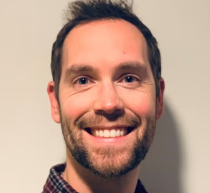 Man with short dark hair and a beard wearing a black and red plaid collared shirt.