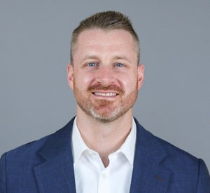 Man with short brown hair and a beard wearing a navy blue suit jacket over a white collared shirt.