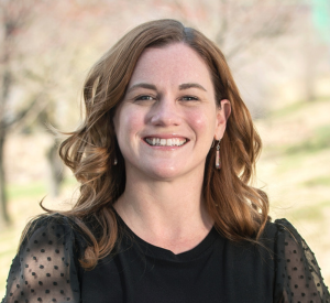 Woman with curly auburn hair wearing a black blouse with sheer sleeves.