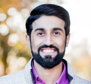 A man with black hair and a beard wearing a gray jacket over a pink collared shirt.