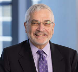 Man with white hair and facial hair wearing glasses and a black suit jacket over a light purple collared shirt and a purple and white patterned tie.