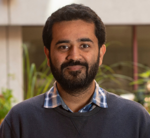 Man with short black hair and facial hair wearing a dark gray sweater over a blue and tan plaid collared shirt.