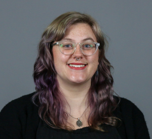 Woman with light brow and purple dyed hair wearing clear glasses and a black sweater.